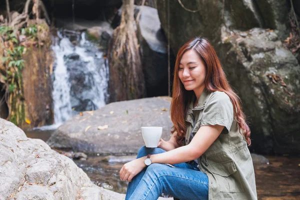 Güzel bir Asyalı kadın şelalenin önündeki kayanın üzerinde oturmuş kahve içiyor.