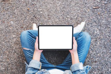 Üst görüntüde, elinde siyah tablet olan beyaz bir kadın açık havada otururken görüntüsü var. 