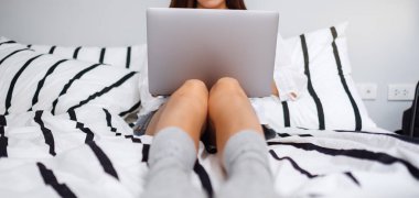 Evdeki rahat beyaz bir yatakta otururken dizüstü bilgisayar kullanan ve çalışan bir kadının yakın plan görüntüsü.