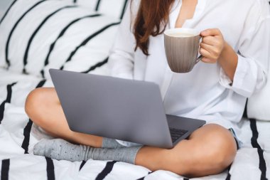 Laptop bilgisayar kullanan ve çalışan bir kadının yakın plan görüntüsü. Evdeki rahat beyaz bir yatakta oturup kahve içen bir kadın.