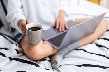 Laptop bilgisayar kullanan ve çalışan bir kadının yakın plan görüntüsü. Evdeki rahat beyaz bir yatakta oturup kahve içen bir kadın.