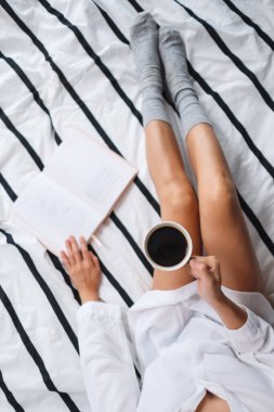 Kitap okuyan ve sıcak kahve içen bir kadının sabahları evdeki rahat bir yatakta görüntüsü.