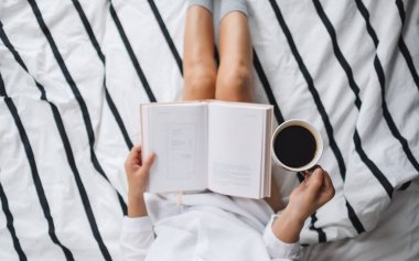 Kitap okuyan ve sıcak kahve içen bir kadının sabahları evdeki rahat bir yatakta görüntüsü.