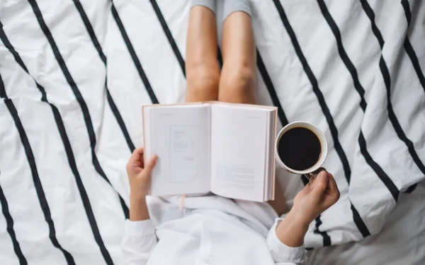 トップ表示画像の女性読書本と飲み物熱いコーヒーで白い居心地の良いベッドで自宅で午前中 — ストック写真