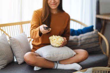 Close up image of a beautiful young woman eating pop corn and searching channel with remote control to watch tv while sitting on sofa at home