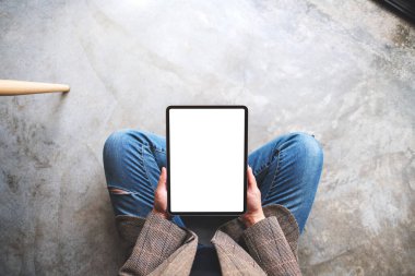 Yerde otururken boş beyaz masaüstü ekranlı tablet bilgisayarını tutan ve kullanan bir kadının en üst görünüm görüntüsü 