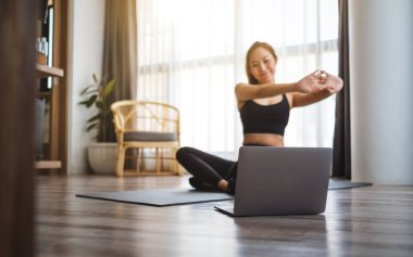 A beautiful young asian woman stretching arms while watching online workout tutorials on laptop at home