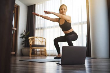 Güzel bir Asyalı kadın evde dizüstü bilgisayar başında antrenman yaparken çömeliyor.