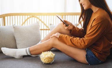 Close up image of a beautiful young woman using mobile phone with a bowl of pop corn on sofa 