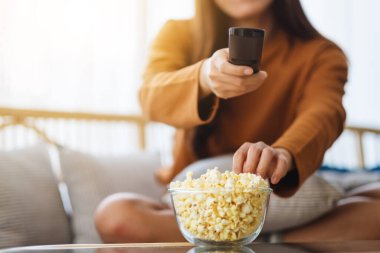 Close up image of a beautiful young woman eating pop corn and searching channel with remote control to watch tv while sitting on sofa at home
