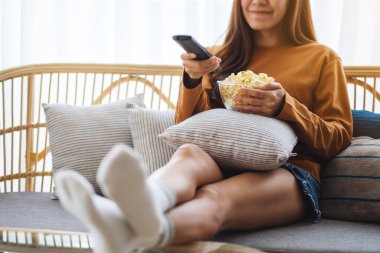 Close up image of a beautiful young woman eating pop corn and searching channel with remote control to watch tv while sitting on sofa at home