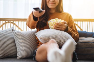 Close up image of a beautiful young woman eating pop corn and searching channel with remote control to watch tv while sitting on sofa at home