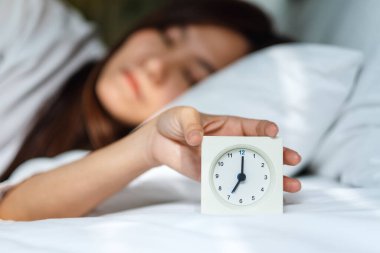 Asyalı bir kadın sabahın köründe beyaz bir yatakta uyurken çalar saati kapatıyor.