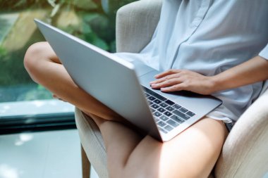 Evdeki yeşil arka planlı beyaz koltukta oturan bir kadının dizüstü bilgisayarda çalışırken çekilmiş yakın plan görüntüsü.