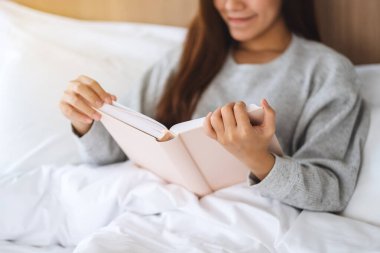 Closeup image of a beautiful young woman reading book in a white cozy bed at home