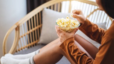 Close up image of a beautiful young woman holding and eating pop corn while sitting on sofa at home