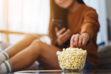 Close up image of a young woman eating pop corn while using mobile phone on sofa at home
