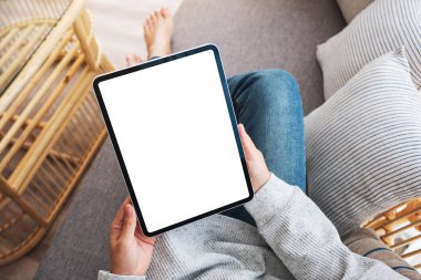Üst görünümde, evde bir kanepede yatarken elinde boş masaüstü beyaz ekranı olan siyah tablet bir kadının görüntüsü var.