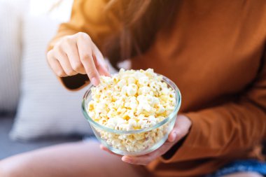 Close up image of a woman holding and picking pop corn from a bowl to eat while sitting on sofa at home