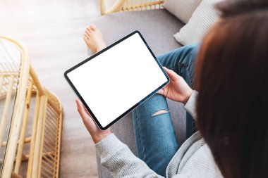 Üst görünümde, evde bir kanepede yatarken elinde boş masaüstü beyaz ekranı olan siyah tablet bir kadının görüntüsü var.