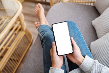 Üst görünümde, evde bir kanepede yatarken elinde boş masaüstü beyaz ekranı olan cep telefonu tutan bir kadının görüntüsü var.