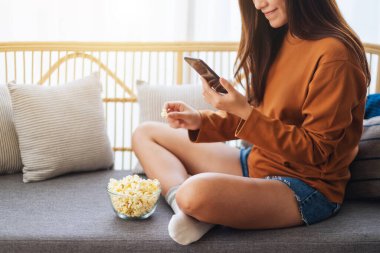 Close up image of a beautiful young woman using mobile phone with a bowl of popcorn on sofa 