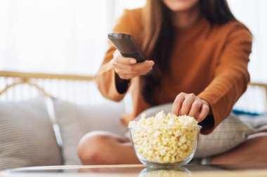 Close up image of a young woman eating popcorn and searching channel with remote control to watch tv while sitting on sofa at home