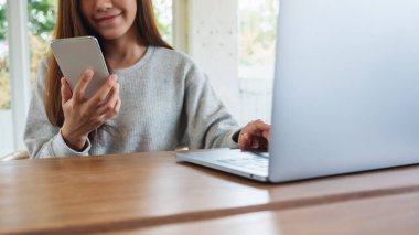 Güzel bir genç kadın cep telefonunu ahşap masada dizüstü bilgisayarla tutuyor ve kullanıyor.