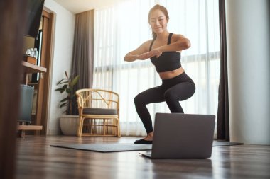 Güzel bir Asyalı kadın evde dizüstü bilgisayar başında antrenman yaparken çömeliyor.