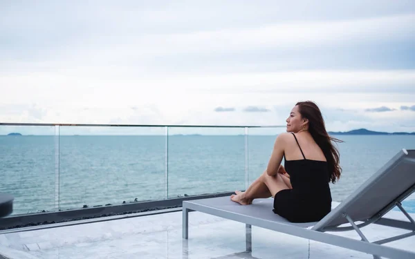 一位年轻美丽的亚洲女子坐在天台游泳池旁边 望着大海和蓝天的景色 — 图库照片
