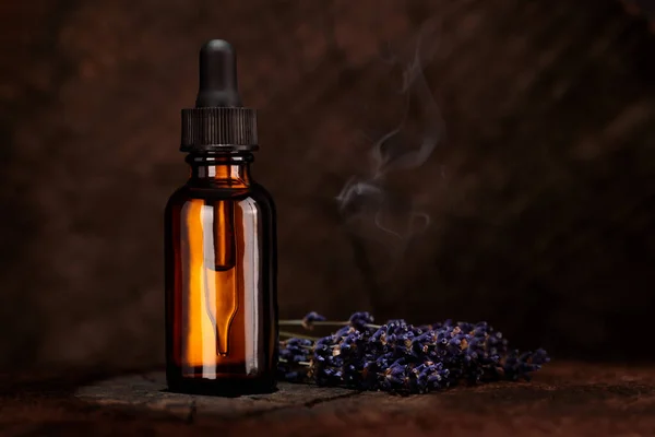 A bottle of cosmetic oil stands on a wooden background. Purple lavender flowers lie on the table. Mock-up of a cosmetic product.
