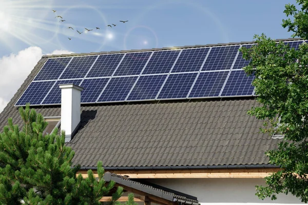 Solar panels on rooftop against blue sky and sun. Solar panels on the roof with a reflective sun and lens. House surrounded by trees, nature.