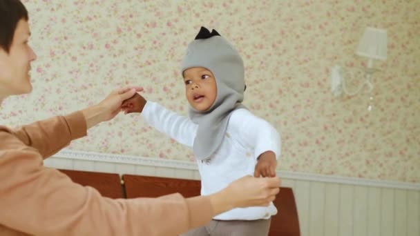 Madre cariñosa muestra un lindo sombrero de dinosaurio a su lindo hijo afroamericano, saltando en la cama, familia amigable preparándose para un paseo, eligiendo ropa juntos, educando a la psicología — Vídeo de stock