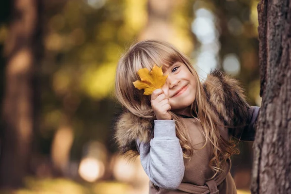 Hermosa chica rubia cubre un ojo con hoja de arce amarillo al aire libre en el parque de otoño. — Foto de Stock