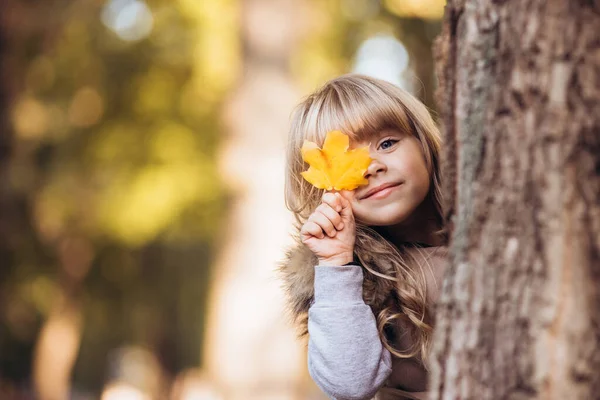 Mooi blond meisje bedekt een oog met gele esdoorn blad buiten in de herfst park. — Stockfoto
