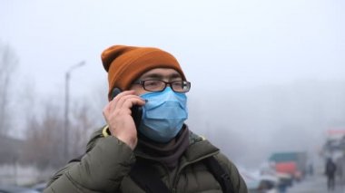 Koruyucu tıbbi maskeli bir adam sokağın ortasında dikilip etrafa bakıyor ve akıllı telefonuyla konuşuyor..