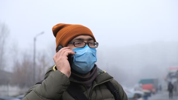 Мужчина в защитной медицинской маске стоит посреди улицы, оглядываясь и разговаривая на своем смартфоне. — стоковое видео