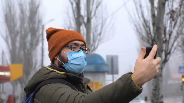 Мужчина в защитной медицинской маске стоит посреди улицы и фотографирует себя на смартфоне. — стоковое видео