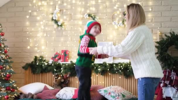 Menino alegre em um chapéu de elfo pula na cama segurando as mãos de suas mães — Vídeo de Stock