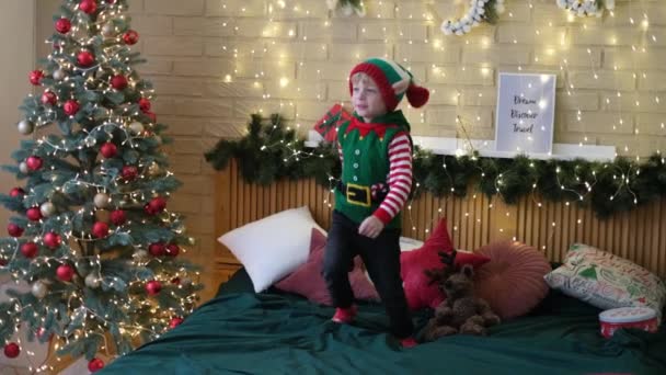 Menino alegre em um chapéu de elfo pulando na cama com uma bengala inflável em suas mãos — Vídeo de Stock