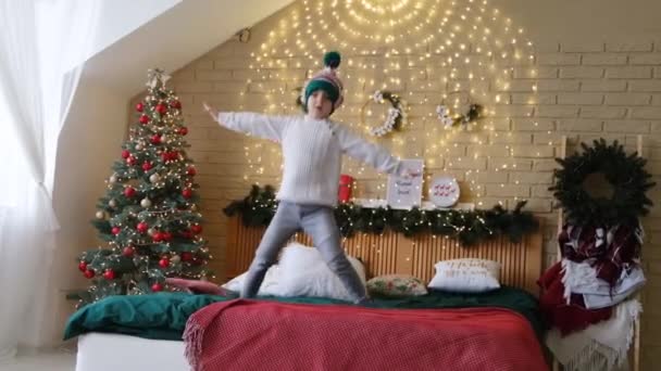 Маленький веселий хлопчик в ельфійському капелюсі стрибає на ліжку з надувною тростиною в руках — стокове відео