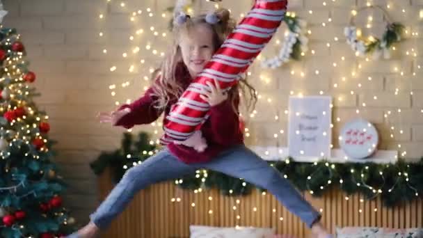 Petite fille joyeuse sautant sur le lit près d'un arbre de Noël élégant avec une canne gonflable dans ses mains — Video