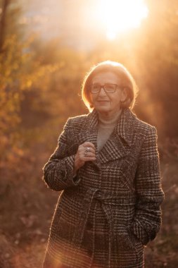 Yünlü ceketli, güzel ve zayıf yaşlı bir kadın, eşarp poz veriyor, sonbahar parkında altın yapraklarla güzel bir ağacın altında duruyor, dinleniyor ve mutlu emekliliğin tadını çıkarıyor.