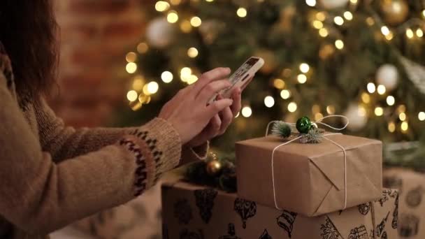 Buying New Years gifts over the Internet. Womens hands with a smartphone in their hands on the background of a Christmas tree. addiction to social networks and chats. Modern holidays online — Vídeo de Stock