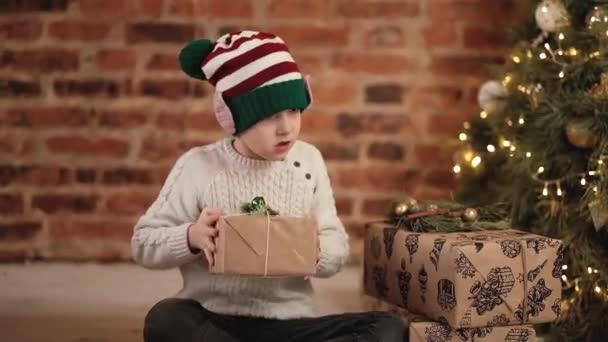 A curious boy takes Christmas presents from under the tree, shakes it, tries to find out whats inside. Happy christmas for baby — Vídeo de Stock