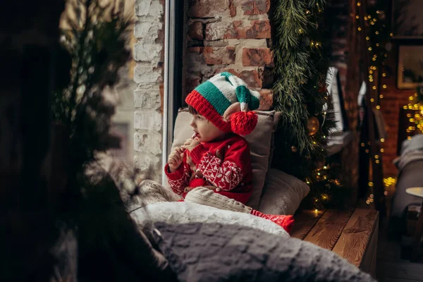 Petit garçon dans un chapeau d'elfe avec un bonbon à la main assis à la fenêtre. Enfant regarde par la fenêtre et attend Noël, Père Noël — Photo