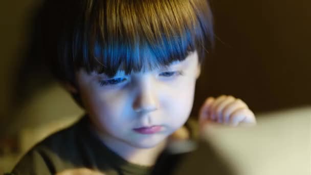 Een schattig jongetje is het kijken naar een tablet scherm terwijl zitten in een kinderstoel op de achtergrond van een kinderkamer. — Stockvideo