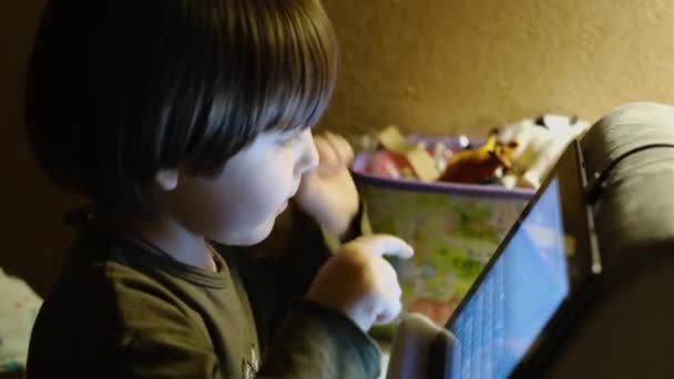 Een schattig jongetje is het kijken naar een tablet scherm terwijl zitten in een kinderstoel op de achtergrond van een kinderkamer. — Stockvideo