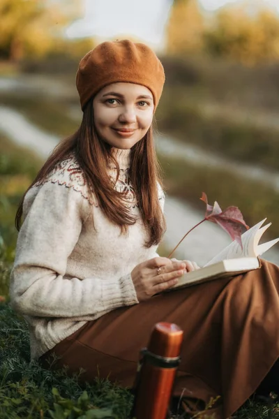 Portrait of a woman in a French beret. A young candid woman in a knitted sweater and brown beret is sitting on the grass reading a book. French girl. Millennial generation.