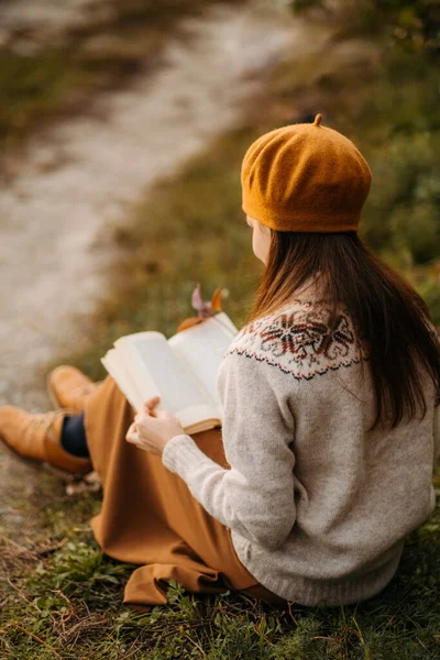 Portrait of a woman in a French beret. A young candid woman in a knitted sweater and brown beret is sitting on the grass reading a book. French girl. Millennial generation.